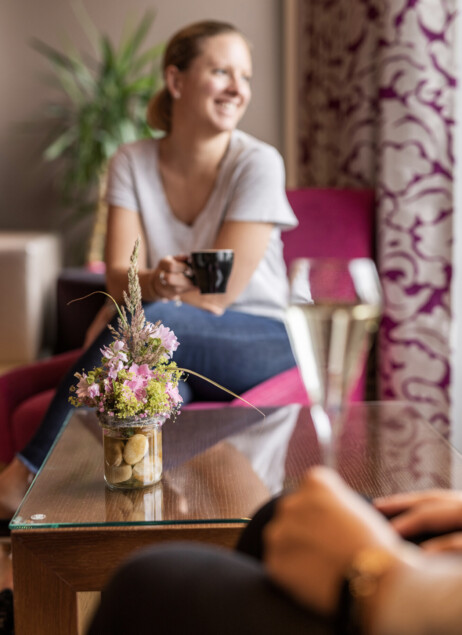 Eine Frau sitzt entspannt mit einer Tasse Kaffee im Seehotel Seewirt Mattsee, im Vordergrund ein Glas mit Blumen.